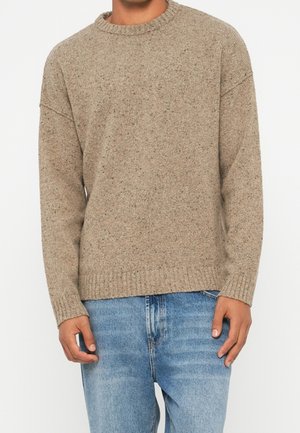 Mann trägt einen beigen Strickpullover mit gerippten Bündchen und Saum, kombiniert mit hellblauen Jeans, steht vor einem schlichten Hintergrund.