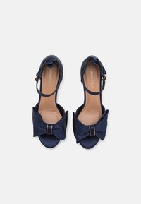 Navy blue high-heeled sandals with a bow accent, open toe, ankle strap, and a smooth texture. Featuring a gold-tone hardware detail.