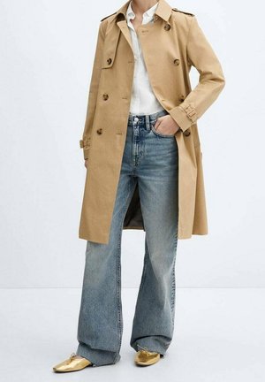 Person wearing beige double-breasted trench coat, white shirt, wide-leg blue jeans, and gold pointed shoes, standing with one hand in pocket.