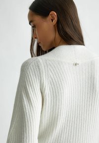 Maglione bianco a coste con una texture morbida, caratterizzato da un sottile dettaglio con chiusura sul collo posteriore.