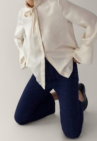 Krämfärgad silkesblus med en rosett vid halsen, knappstängning och vida ärmmuddar; matchad med mörkblå jeans i denim och svarta skor.