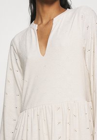 Robe de couleur claire avec un décolleté en V, texture florale complexe, manches longues et détail de taille froncée.