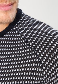 Pull en maille avec col rond, présentant un motif texturé de points alternés marron foncé et blancs. Matière douce et coupe ajustée.