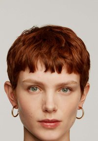 Cabello corto castaño con capas texturizadas, pendientes de aro dorados y un cutis suave con pequeñas imperfecciones visibles.