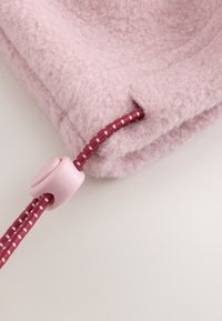 Närbild av ett rosa ludet tyg med en röd- och vitprickig dragsko som trätts genom ett rosa plaststopp.