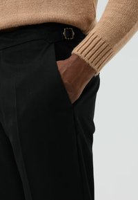 Zwarte op maat gemaakte broek met zijzakken en een gouden gesp aan de tailleband, gecombineerd met een beige gebreide trui met geribbelde manchetten.