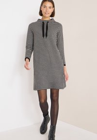 Czarna i biała sukienka w kratę z kapturem, długimi rękawami i sznurkiem przy szyi. Nosi się ją z czarnymi rajstopami i sznurowanymi butami.