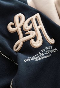 Granatowy sweter z haftowanymi kremowymi literami "LFA", współrzędnymi oraz napisem "LOS ANGELES - CALIFORNIA" w nowoczesnej, czystej czcionce.