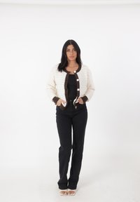 Cárdigan de ganchillo en color crema con ribete marrón, presenta botones grandes y bolsillos. Combinado con jeans acampanados negros y zapatos de punta abierta.