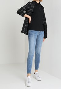 Blazer à carreaux noirs porté sur un polo noir, associé à un jean skinny bleu clair et des baskets blanches. Textures lisses, coupe moderne et style décontracté.