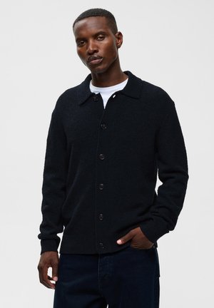 Homme portant un cardigan noir boutonné par-dessus une chemise blanche, la main gauche dans la poche, sur un fond clair uni.