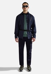 Marineblauwe nylon jas met zwarte riem, gecombineerd met een groene trui, donkere cargobroek en groene sneakers met zwarte accenten en textuurlaces.