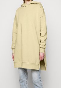 Długa beżowa bluza z kapturem wykonana z miękkiego materiału, wyposażona w kaptur z regulowanym sznurkiem, opadające ramiona oraz boczne rozcięcia, o luźnym kroju.