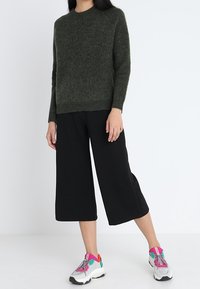 Olivgrüner Strickpullover, schwarze, hochgeschnittene Hosen mit weitem Bein und bunte Sneaker mit pinken und grünen Akzenten. Die Textur wirkt weich und entspannt.