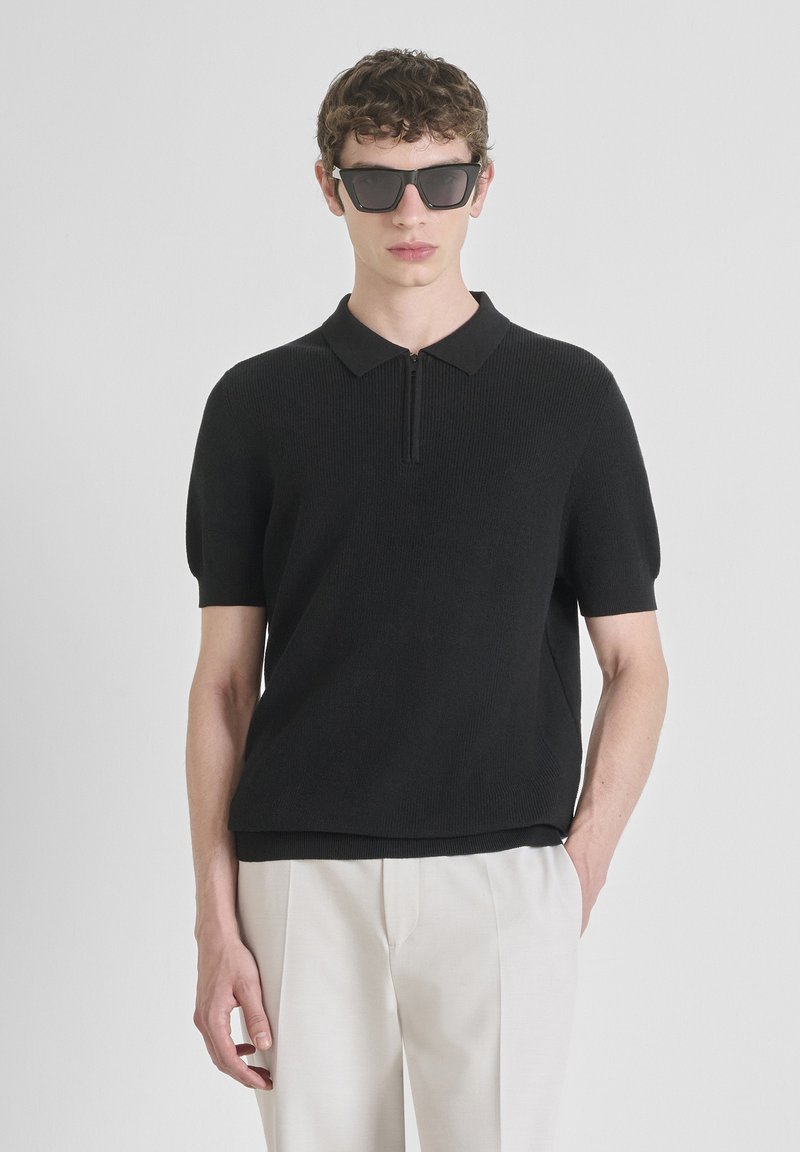 Crna rebrasta polo majica s kratkim rukavima i patentnim zatvaračem. Model nosi pravokutne naočale i svijetle hlače.