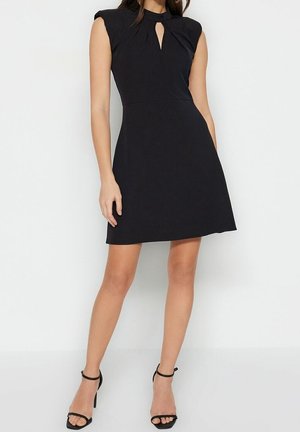 Kvinde iført en sort ærmeløs kjole med nøglehul i halsudskæringen og sorte høje hæle med ankelrem, stående mod en hvid baggrund.