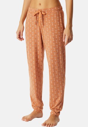 Frau trägt orange Schlafanzughose mit weißen Blumenmustern und gebundenem Bund, steht barfuß vor schlichtem Hintergrund.