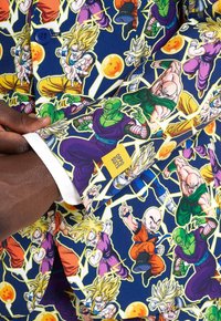 Farbenfrohe Anzugjacke aus blauem Stoff, geschmückt mit lebhaften Dragon Ball-Charakteren, umrandet in Gelb, mit einem weißen Manschetten-Detail und einem Logosticker.