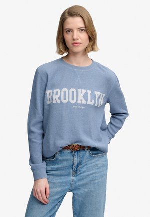 Kvinde med kort blondt hår iført en lyseblå sweater med teksten "BROOKLYN" og blå jeans, stående med den ene hånd på hoften.