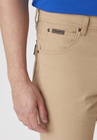 Mannens vänstra hand vid beige Wrangler-byxans ficka, iförd en blå skjorta, närbild som visar tygtexturen och sömnadsdetaljer.