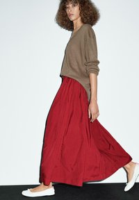Pull en maille marron avec des épaules tombantes et un col en V profond, associé à une jupe maxi rouge fluide et des chaussures plates blanches.
