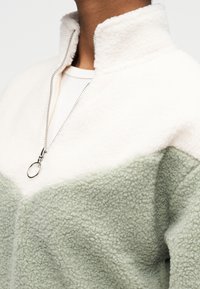 Φλις πουλόβερ με ψηλό γιακά. Διαθέτει εκρού πάνω μέρος και πράσινο κάτω μέρος. Ασημένιο φερμουάρ και λεπτομέρειες με υφή στην επιφάνεια.