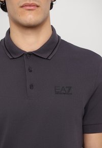 Tmavo šedá polo košeľa vyrobená z texturovanej tkaniny, s klasickým golierom, trojbodovým zapínaním a čiernym vyšitým logom na hrudi.