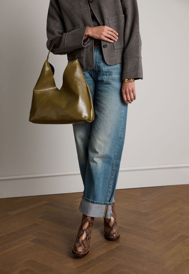 Borsa a mano in pelle verde oliva con un design triangolare, jeans in denim blu rilassati e stivali alla caviglia in pitone marrone con finitura lucida.