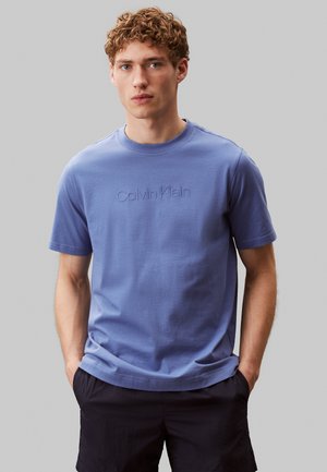 Giovane uomo con capelli ricci che indossa una maglietta blu Calvin Klein e pantaloncini neri, in piedi con le mani nelle tasche davanti a uno sfondo neutro.