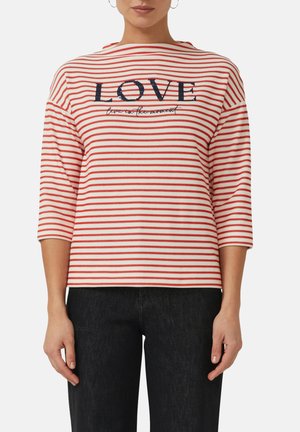 Femme portant un chemisier rayé rouge et blanc avec le mot « LOVE » et un texte en écriture cursive sur la poitrine, associé à un pantalon noir.