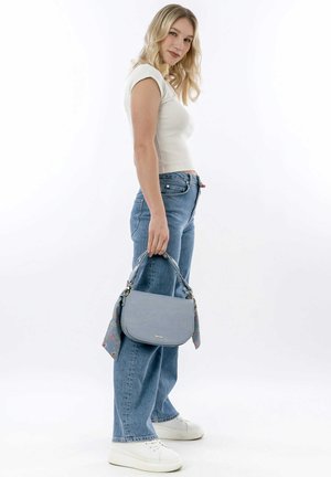 Junge Frau in weißem Oberteil und Jeans hält eine blaue Handtasche mit einem floralen Schal und steht vor weißem Hintergrund.