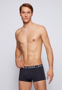 Schwarze Boxershorts mit einem elastischen, markierten Bund, auf dem "HUGO BOSS" in Weiß steht. Glattes Material und körpernaher Schnitt. Das Modell ist oberkörperfrei.