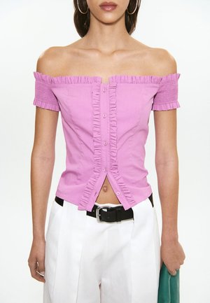 Femme portant un haut rose à épaules dénudées, volanté et boutonné, associé à un pantalon blanc taille haute et une ceinture noire, tenant une pochette verte.