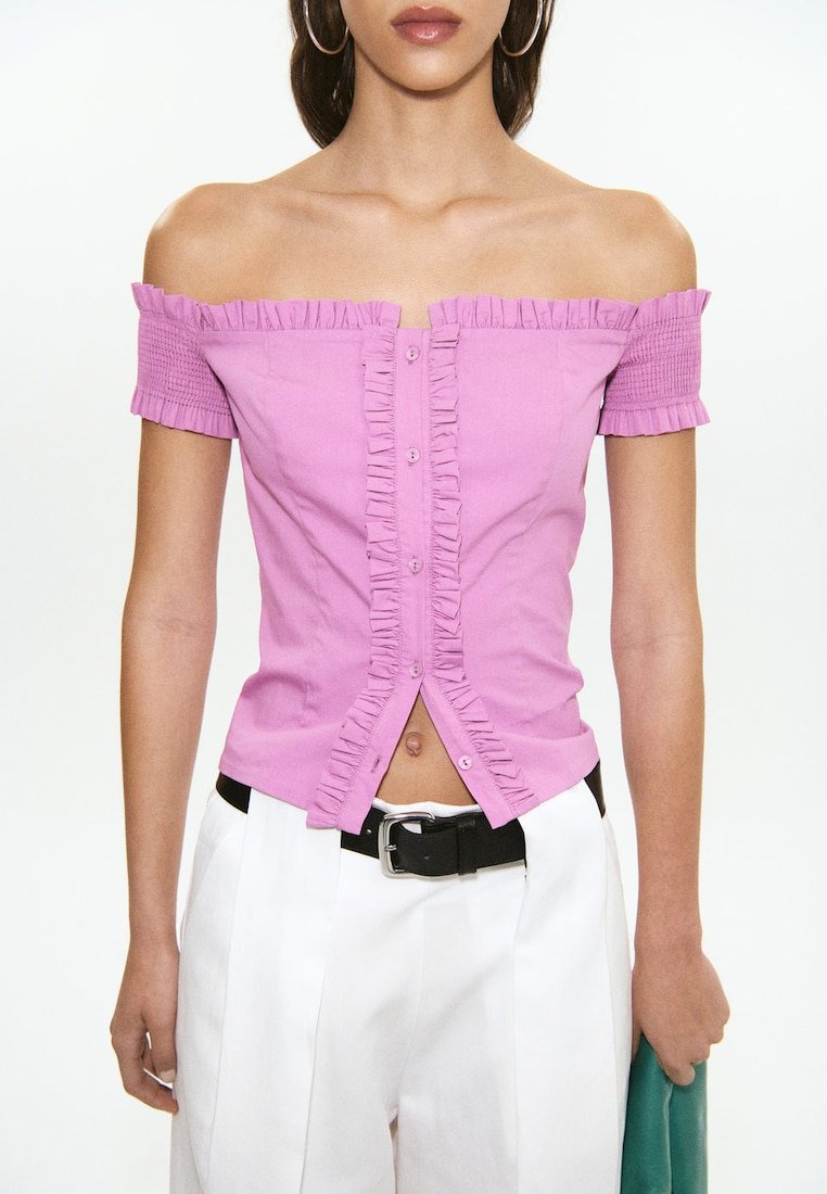 Femme portant un haut rose à épaules dénudées, volanté et boutonné, associé à un pantalon blanc taille haute et une ceinture noire, tenant une pochette verte.