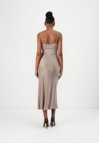 Abito midi in raso beige con spalline sottili e silhouette aderente, dotato di una chiusura sul retro discreta e una texture liscia. Abbinato a tacchi neri.