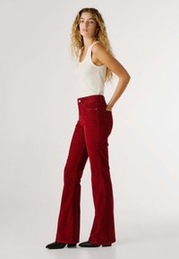 Pantaloni a zampa d'elefante in corduroy rosso con tasche frontali, vita alta e una trama sottile. Abbinati a una maglietta bianca senza maniche e scarpe nere con punta.
