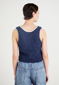 Mørkeblå, ermeløs topp med teksturert stoff; har en hvit scalloped kant ved halsen og en cropped, avslappet passform.