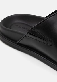 Sorte skinnslippers med glatt tekstur, buet form, søm i siden, og en polstret såle med preget merke.