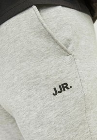 Gros plan sur la poche d'un pantalon de survêtement gris clair avec le logo noir "JJR." imprimé près de la couture, porté avec un haut foncé.
