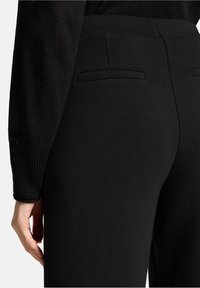 Czarne elastyczne spodnie o gładkiej fakturze, z dwoma tylnymi kieszeniami i dopasowanym krojem. Widoczna ręka dotyka tkaniny.