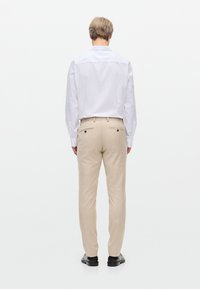 Pantaloni beige con una vestibilità su misura, dotati di due tasche posteriori e dettagli di cuciture sottili, abbinati a una camicia bianca a maniche lunghe.