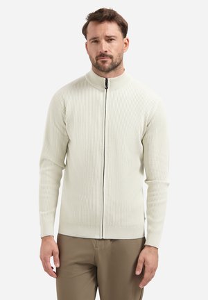 Mann in einem cremefarbenen, gerippten Full-Zip-Pullover mit hochgeschlossenem Kragen und braunen Hosen, vor einem schlichten weißen Hintergrund stehend.