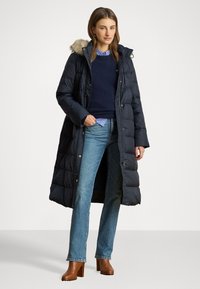 Tengerészkék puffasztott kabát, prémes kapucnival, gombos záródással és steppelt kialakítással. Tengerészkék pulóver és kék farmer fölött viselve. Barna magassarkú cipő.