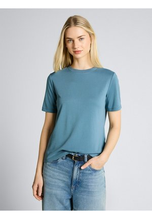 Blondynka z długimi włosami, ubrana w turkusową koszulkę z krótkim rękawem, jasnoniebieskie dżinsy i czarny pasek, stoi z jedną ręką w kieszeni.