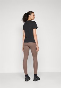 Kvinne står med ryggen til iført en svart t-skjorte, brune leggings, svarte sokker og svarte joggesko mot en enkel bakgrunn.