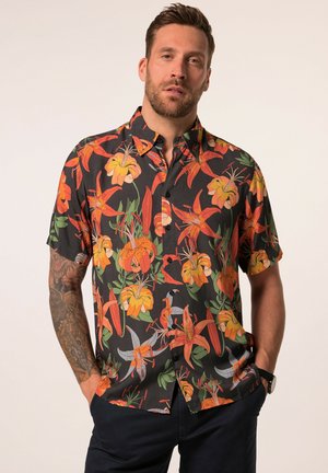 Uomo che indossa una camicia nera a maniche corte con grandi motivi di fiori di giglio arancioni e gialli, in piedi con le mani in tasca.