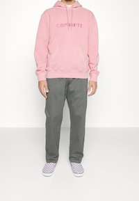 Růžová mikina s klokánkovým kapsou a vyšívaným logem, doplněná o šedé kalhoty a fialové kostkované slip-on boty.