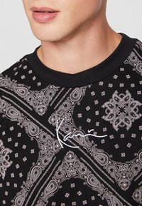T-shirt nera con motivo paisley bianco, con collezione rotonda e disegni intricati. Include un logo ricamato bianco al centro.
