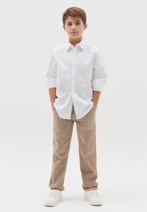 Witte lange mouwen blouse met knoopsluiting, beige broek en witte sneakers. De blouse heeft een klassieke kraag, de broek heeft zijzakken en een relaxte pasvorm.