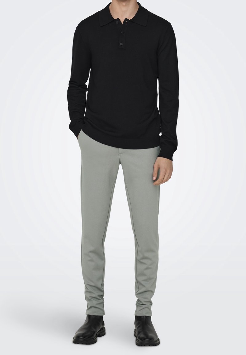 Homme portant un polo noir à manches longues, un pantalon slim gris clair et des bottes en cuir noires, debout les mains dans les poches.